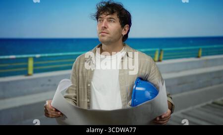 Man holding blueprints in promenade setting, wearing hardhat by the sea, focused on construction planning. Stock Photo