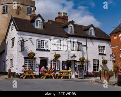 Ludlow, UK - Aug 09 2025: Tourists relax at picnic tables outside The Wheatsheaf Inn on a bright summer day in Ludlow, Shropshire, UK. Historic pub facade with hanging baskets and blue sky. Stock Photo