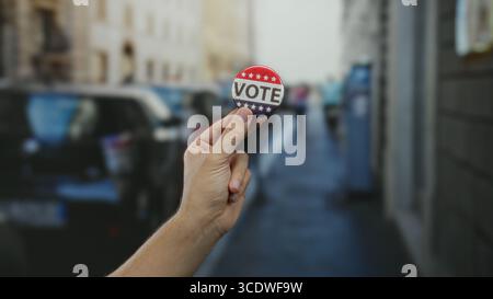 Man holding vote button on street showcasing election engagement outdoors with city backdrop and caucasian hand emphasizing political participation in Stock Photo