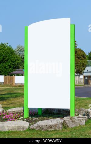 A large blank signboard with bright green edges stands on grass surrounded by rocks and pink flowers. Stock Photo