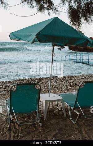 Beach scene at sunset with turquoise umbrella and sun loungers, small table between loungers, sea with small waves Stock Photo