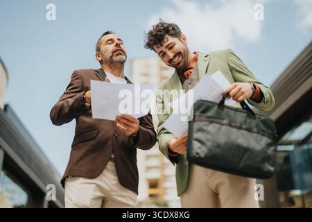 Two businessmen laughing while discussing documents outdoors on a sunny day Stock Photo