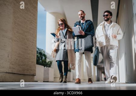 Three colleagues walking outdoors while discussing work and carrying documents Stock Photo