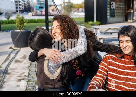 Three cheerful young women embrace at a table outside a cafe, enjoying friendship and connection Stock Photo