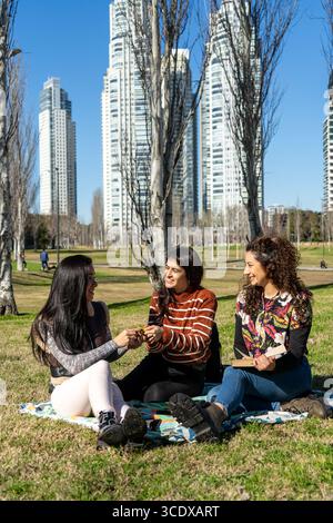 Three young women enjoy a sunny day in a city park, sharing flowers and laughter while sitting on a blanket Stock Photo
