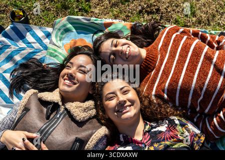 Three cheerful young women relaxing together on a blanket in a park, enjoying a sunny day and each other's company Stock Photo