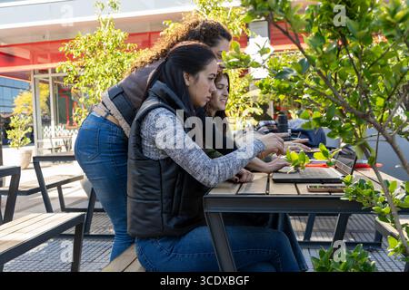 Three businesswomen collaborating on a project, working together on a laptop in a modern outdoor urban setting Stock Photo