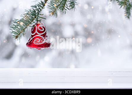 Christmas background with empty white wooden board surface for product placement, red bell ornament hanging from evergreen branch covered in snow Stock Photo