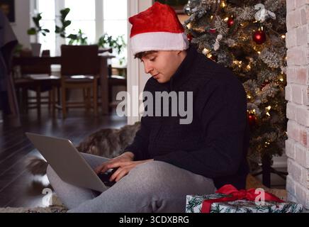 Young man in Santa hat sitting under Christmas tree using laptop computer Stock Photo