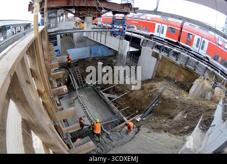 Construction at Münster railway station with workers installing rebar and pouring concrete for an underground structure. A red regional train passes a Stock Photo