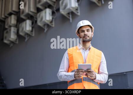 Construction engineer in safety gear inspects a building's air conditioning systems with a tablet. Stock Photo