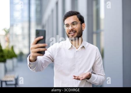 Smiling man holding smartphone and engaging in an interactive way outside a modern building. Stock Photo