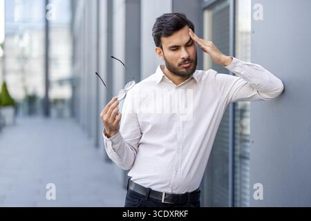 A young man leans against a wall holding his glasses, deep in thought, outdoors. Stock Photo