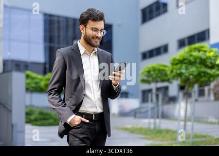 A confident man dressed in business attire, engaged, smiling as he checks his smartphone outside a modern office building, exhibiting productivity, connectivity, and professionalism. Stock Photo