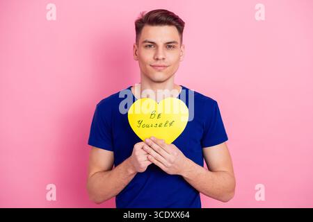 Young man holding yellow heart sign with positive message, standing against pink background wearing casual blue shirt Stock Photo
