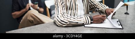 Header of Black man signing document at counter in bank office while anonymous Caucasian man sitting in background using smartphone, both engaged in separate financial activities Stock Photo