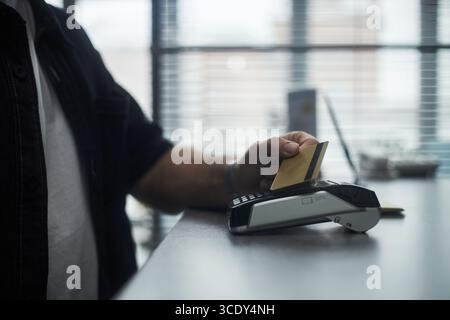 Anonymous man holding credit card making contactless payment using point of sale terminal at bank office counter, partial view showing hand and device interaction Stock Photo