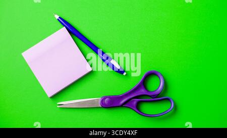 A set of stationery laid out on a bright green surface features a pair of purple scissors, a blue pen, and a stack of sticky notes. These tools are id Stock Photo