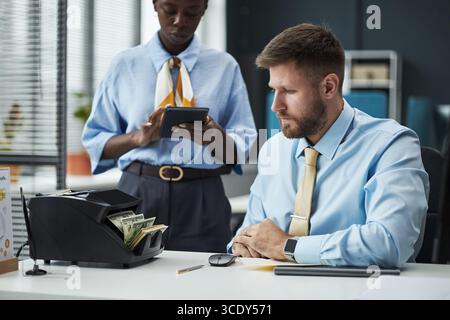Caucasian man sitting at desk operating money counting machine while Black young adult woman standing nearby using digital tablet in modern bank office environment Stock Photo