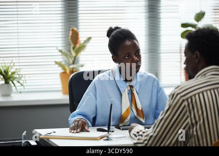 Young Black female bank assistant sitting at desk talking to middle aged man in office, both engaged in conversation with documents and electronic device on table Stock Photo