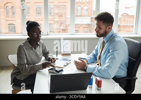 Black young woman making card payment on terminal while sitting next to Caucasian male consultant at desk in bank office, with laptop and paperwork visible Stock Photo