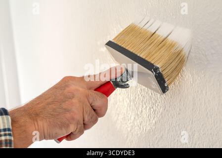 Close-up of a hand. A painter paints a masonry wall with white paint using using the paintbrush. Construction industry. Stock Photo