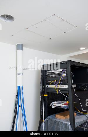 Patched hole in ceiling above Metal server rack with patch panels, firewall appliance and switch with many Cat-6A cables emerging from conduit, USA Stock Photo