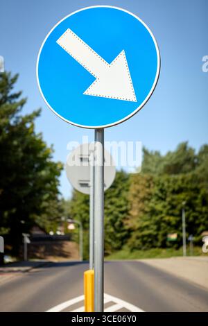 Blue circular keep left road sign with a white arrow pointing downwards to the left, positioned beside a roadway with green trees in the background, i Stock Photo