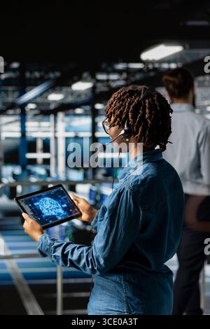 Server hub engineer uses tablet and headset mic to provide remote assistance on industrial platform in data center. Server room professional solves backend issues, coordinating remote teams. Stock Photo