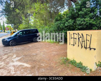 Muro, Spain - Jul 24, 2025: A dark van parked near a yellow wall marked with graffiti reading ISRAEL next to a forested roadside near Playa de Muro, M Stock Photo