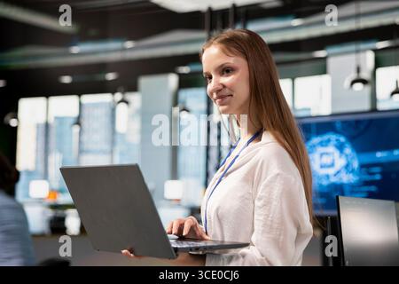 Smiling junior developer utilizing artificial intelligence to optimize performance using laptop. Cheerful young female engineer in office training machine learning models with notebook Stock Photo