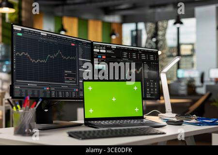 Green screen laptop in brokerage office next to workstation monitor displaying live stock charts. Mockup notebook on desk and computer with financial software in prop firm workspace Stock Photo