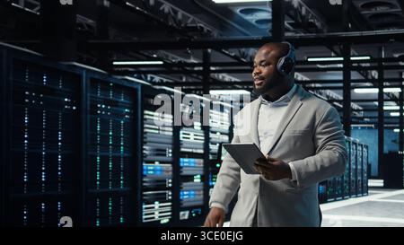 Data center admin wearing headphones using tablet to identify bottlenecks. Server farm IT expert listening music while troubleshooting bugs, implementing optimizations, camera A Stock Photo