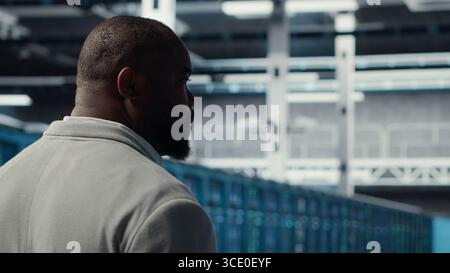 System administrator in data center doing inspection on infrastructure tech handling user requests. African american worker in server farm analyzing traffic patterns of equipment, camera B Stock Photo