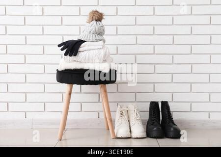 Stack of folded sweaters with hat, gloves on chair and boots near white brick wall Stock Photo