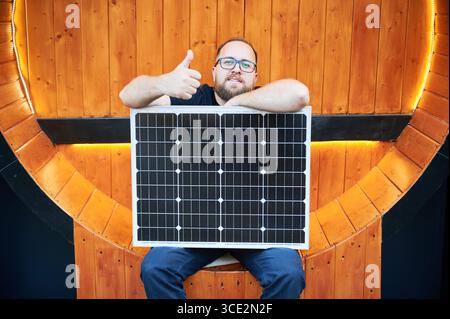 Bearded man happily holding solar panel, giving thumbs up. Guy with glasses sitting against backdrop of warm, wooden planks. Setting appears cozy and modern, with subtle lighting accentuating scene. Stock Photo