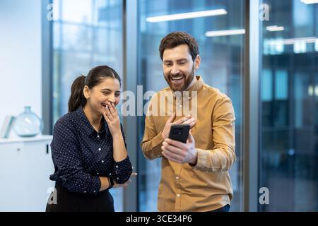 A young woman and a man happily watching something amusing on a smartphone in an office. Stock Photo