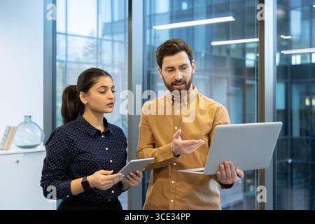 Two colleagues in a modern office setting are collaborating on a project using a laptop and tablet, emphasizing teamwork, technology integration, and professional collaboration in a vibrant workspace Stock Photo