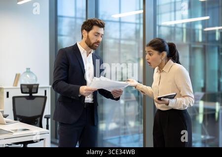 Two office coworkers engage in a lively discussion while reviewing documents and using a tablet in a modern workspace, showcasing collaboration, communication Stock Photo
