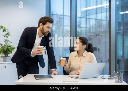 Two coworkers interacting in a contemporary office setting, sharing coffee while discussing business ideas, highlighting teamwork and communication. Stock Photo