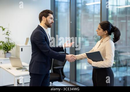 A man and a woman dressed in business attire shaking hands in a bright office setting, creating a professional and cooperative ambiance. Stock Photo