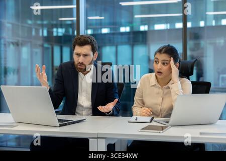Two professionals in a modern office showing confusion as they work on laptops, facing an unexpected issue. The environment reflects a collaborative workspace and professional atmosphere. Stock Photo