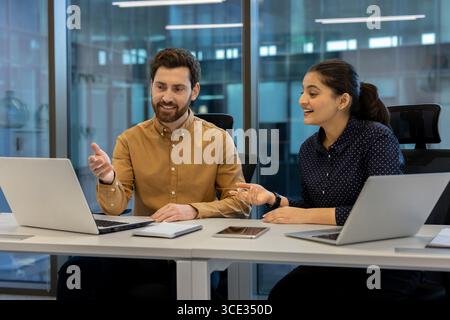 Two coworkers engage in a vibrant discussion while working on laptops in a bright, modern office setting. The image shows teamwork, communication, and productivity in a professional atmosphere. Stock Photo