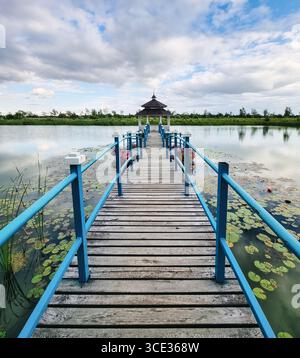 Wooden bridge over a pond leading to a white gazebo. Symmetric diminishing perspective view of a footbridge in countryside on a cloudy day. Stock Photo