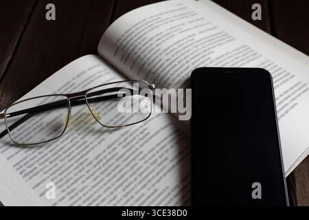 Close-up of eyeglasses and smartphone resting on an open book, symbolizing the balance between traditional reading and modern digital life. Ideal for Stock Photo