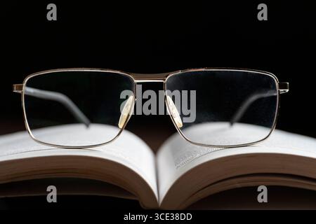 A pair of eyeglasses resting on an open book, symbolizing reading and learning. The dark background enhances focus on the glasses and pages, creating Stock Photo