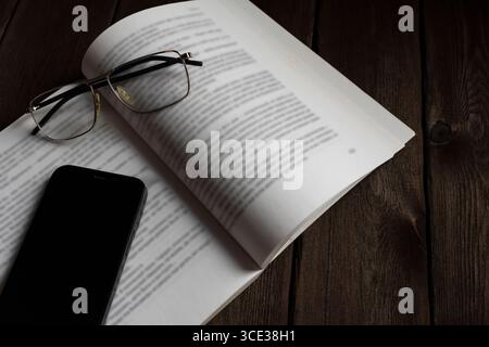 Minimalist close-up of a smartphone and eyeglasses on an open book lying on a dark wooden table. Symbol of learning, distraction, or digital lifestyle Stock Photo