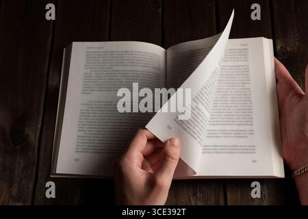 Hands flipping through pages of an open book on a dark wooden table, blurred text highlights the motion of reading and the tactile experience Stock Photo