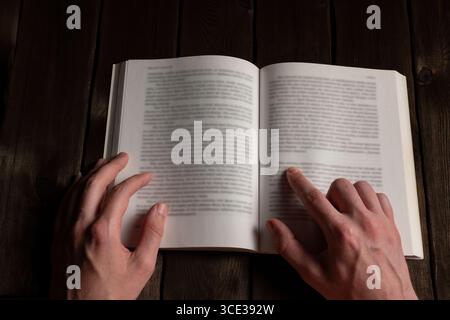 Close-up of hands holding and reading an open book with blurred text, finger pointing at a line. Set on a dark wooden background. Concept of concentra Stock Photo