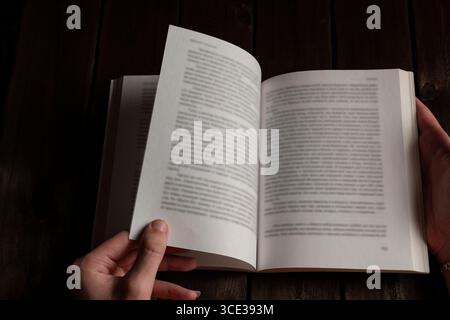 A person turning a page of an open book on a dark wooden table, with hands in focus, capturing a cozy, studious atmosphere perfect for reading and lea Stock Photo